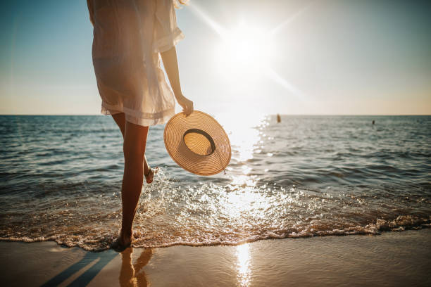 Person in light dress holding sunhat, walking towards ocean at sunset, symbolizing summer attire.