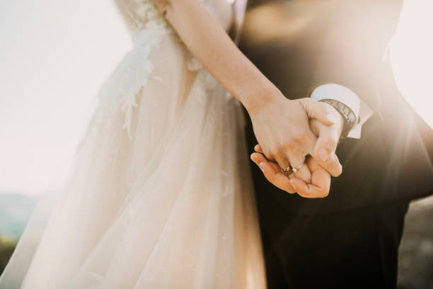 A bride and groom holding hands, showcasing modern wedding attire under a soft sunlight glow.